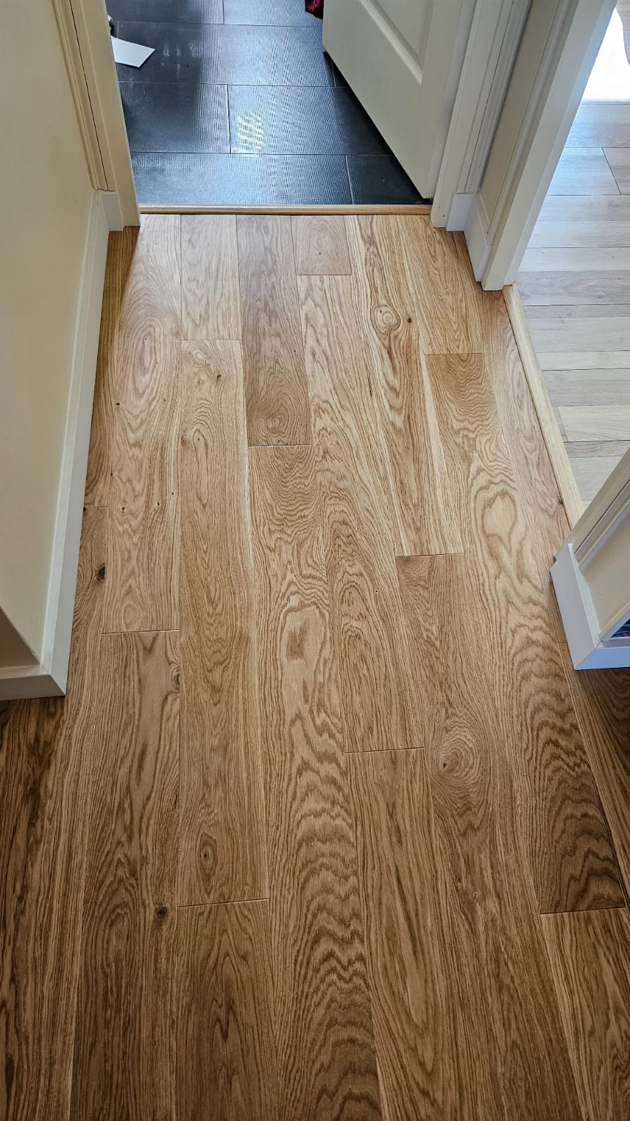 Parquet en chêne clair aux veines apparentes dans un couloir avec plinthes blanches et seuils de porte.