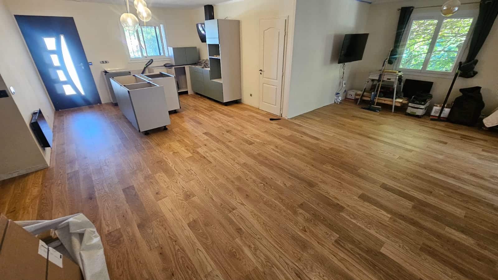 Pose de parquet en chêne clair dans une cuisine en rénovation avec meubles gris et porte d'entrée bleue moderne.