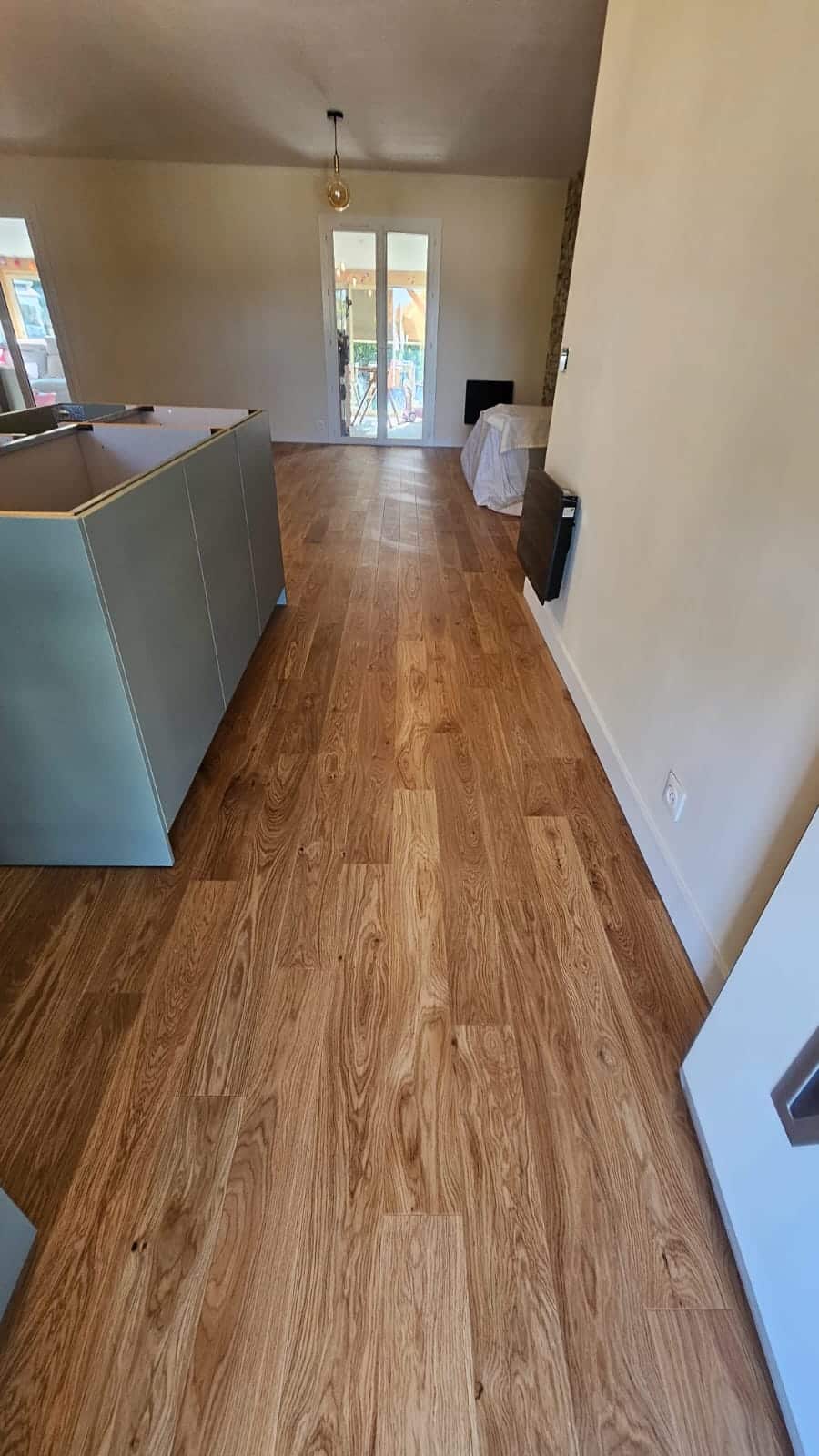 Parquet en chêne clair posé dans une cuisine moderne lumineuse avec îlot central gris et grandes fenêtres au fond.