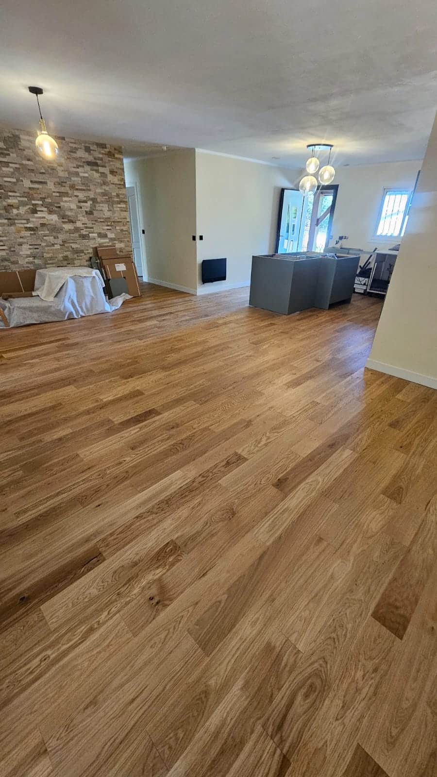 Salon lumineux avec parquet chêne clair, mur en pierre et îlot central gris dans une maison moderne rénovée.
