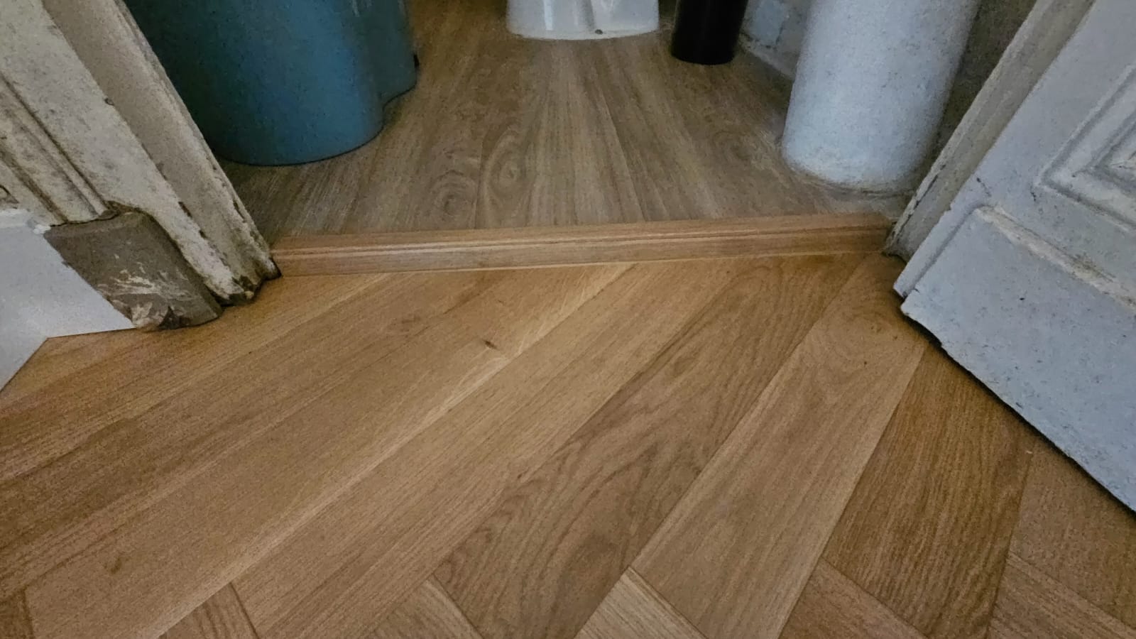 Transition entre parquet en chêne en chevrons et sol stratifié avec barre de seuil en bois dans un encadrement de porte.