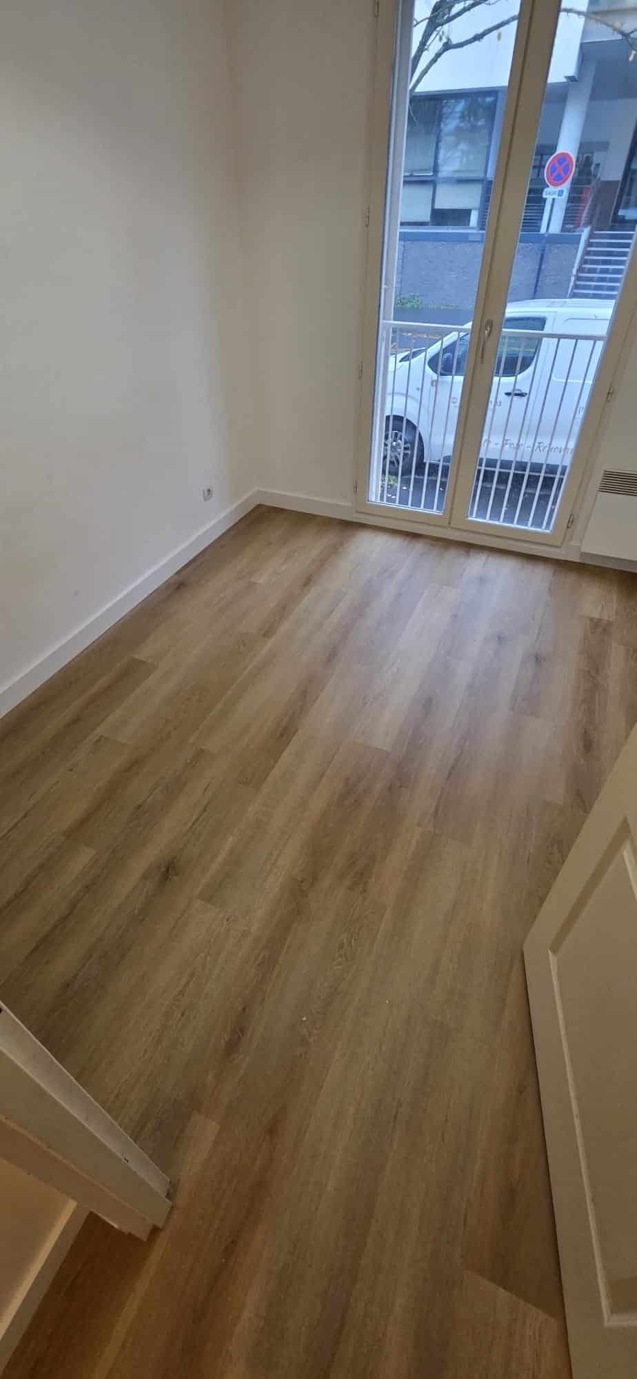 Pièce lumineuse avec parquet stratifié en bois clair, murs blancs et grande porte-fenêtre donnant sur l'extérieur.