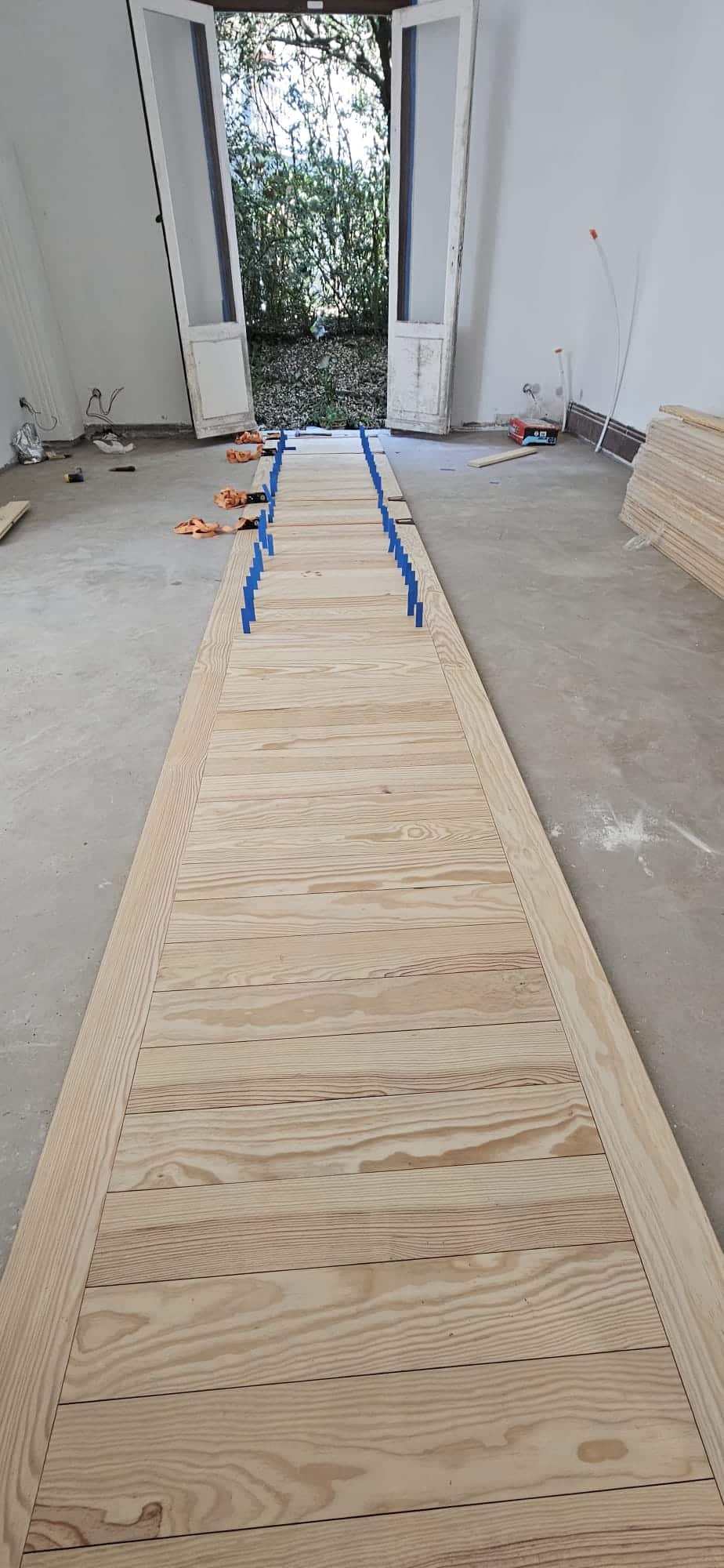 Installation d'un parquet en bois clair dans une pièce en rénovation avec des cales bleues et vue sur le jardin.