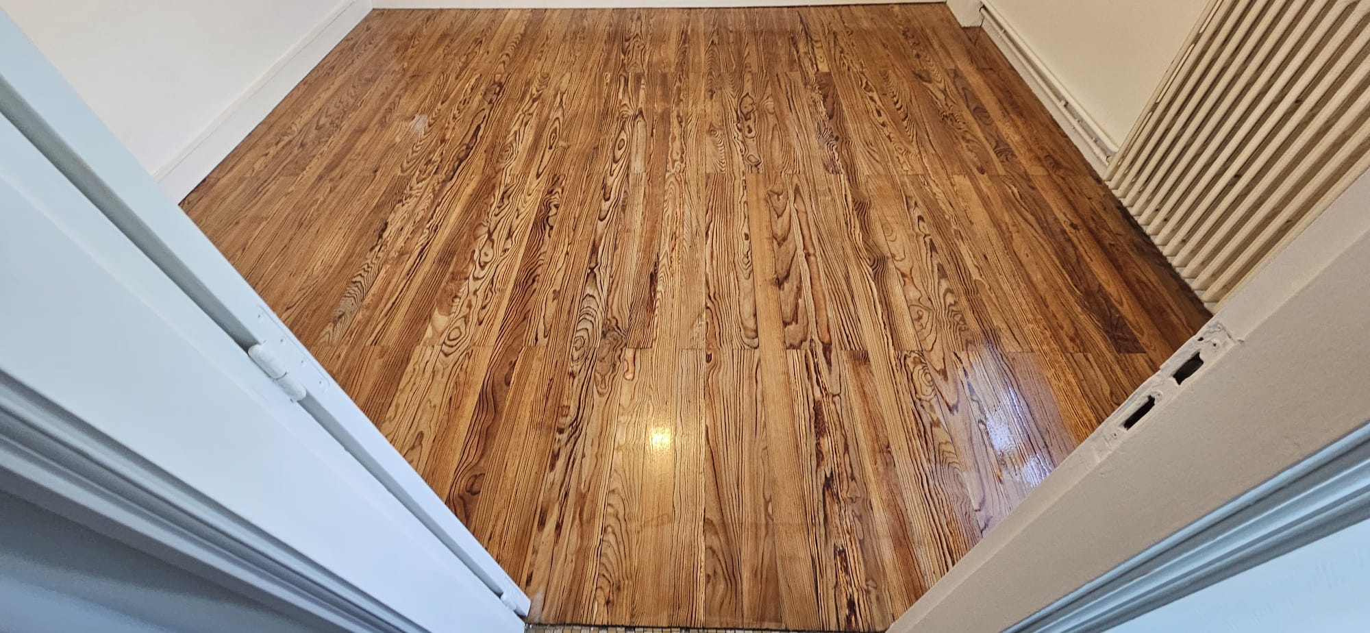 Parquet en Bois Massif Brillant : Rénovation Intérieure Parquet en bois massif à finition brillante et veinures marquées dans une pièce vide aux murs blancs.