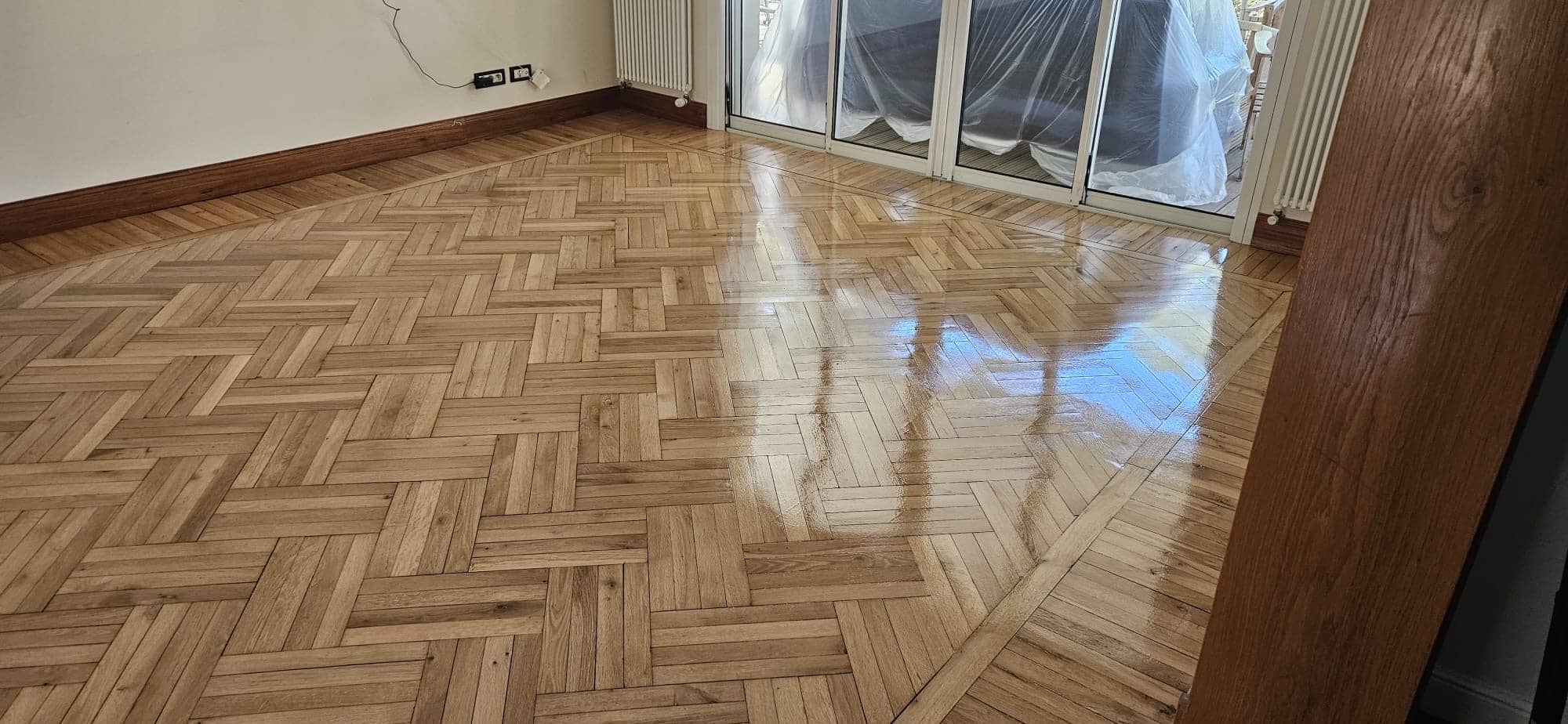Parquet en bois motif vannerie fraîchement verni et brillant dans une pièce vide lumineuse avec plinthes sombres.