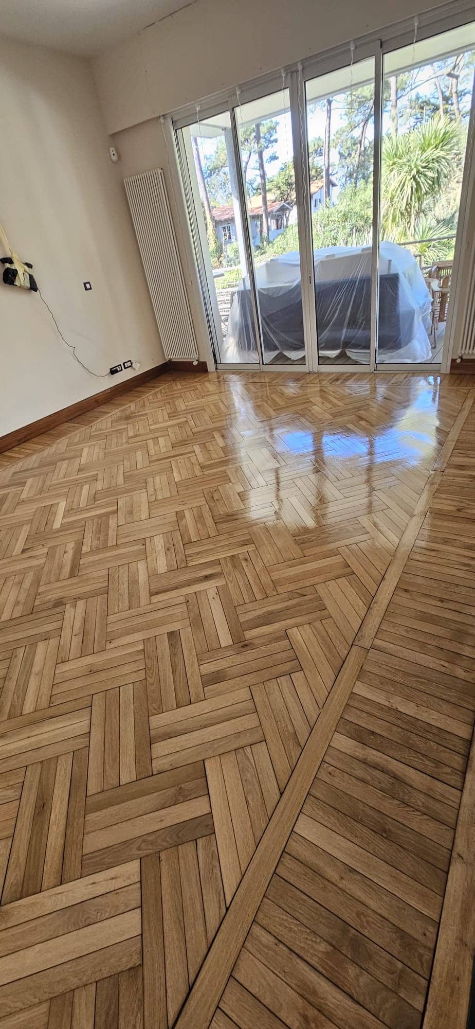 Parquet en chêne massif au motif complexe, vitrifié et brillant, dans un salon lumineux avec baie vitrée.