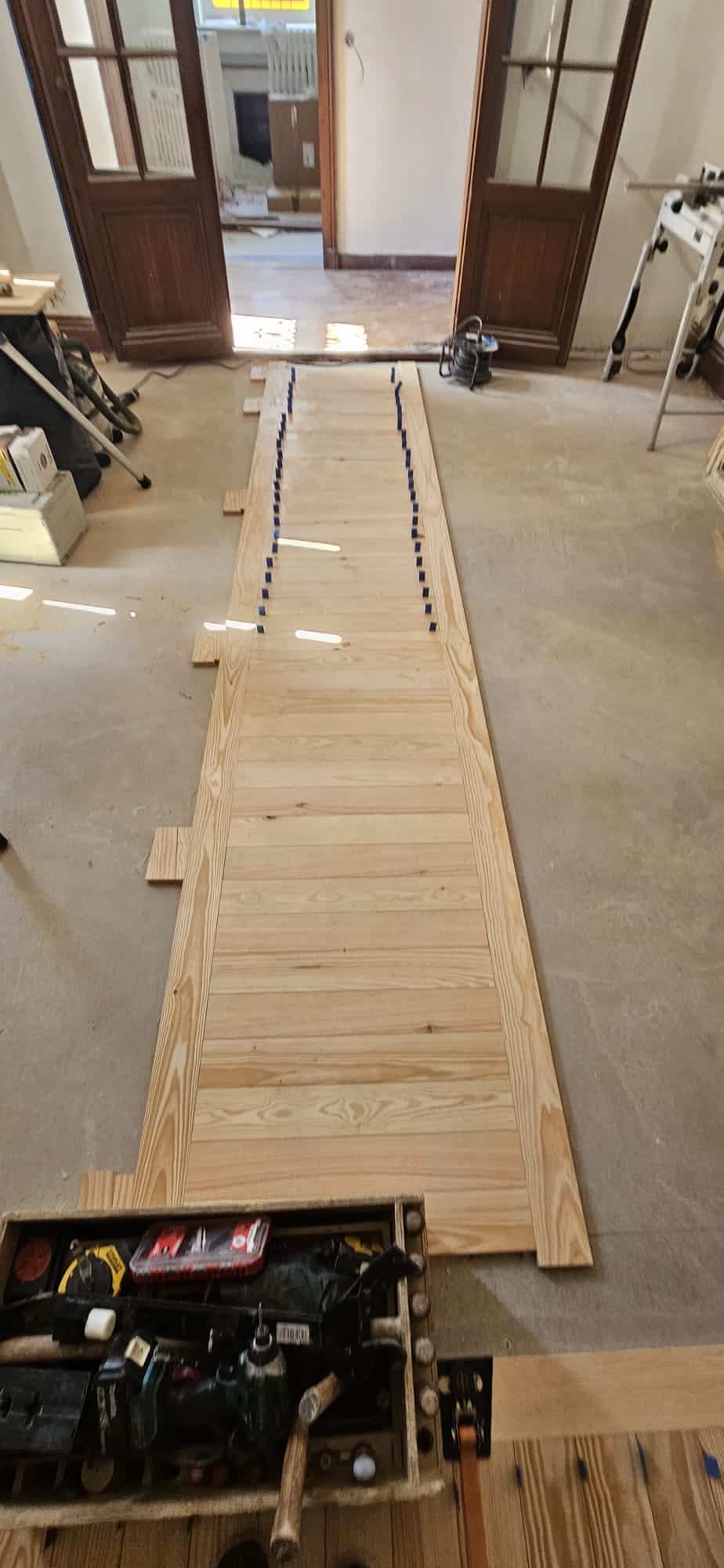 Pose de plancher en bois massif clair avec outils de menuiserie et cales bleues dans un chantier de rénovation.