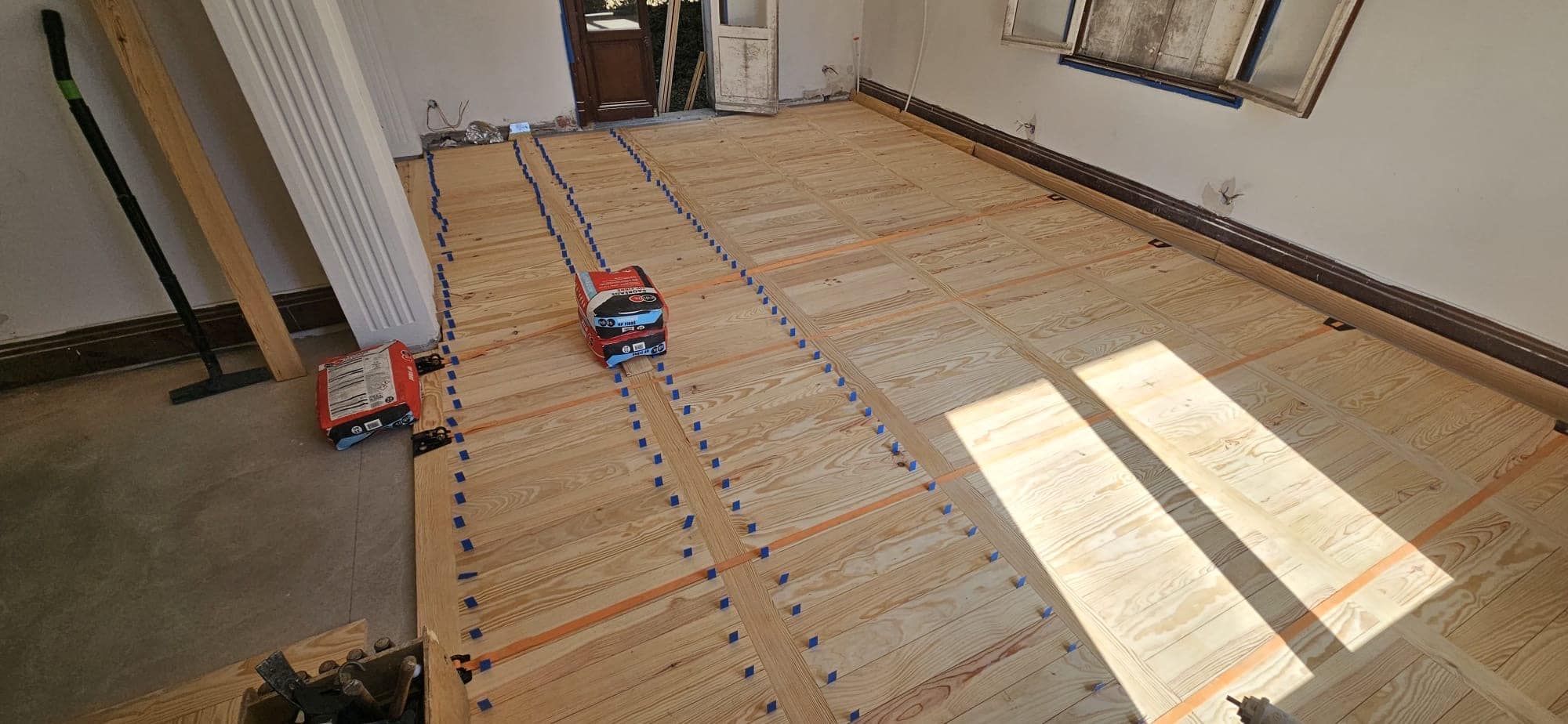 Pose de parquet en pin massif avec cales bleues dans une pièce en rénovation avec sacs de mortier au sol.