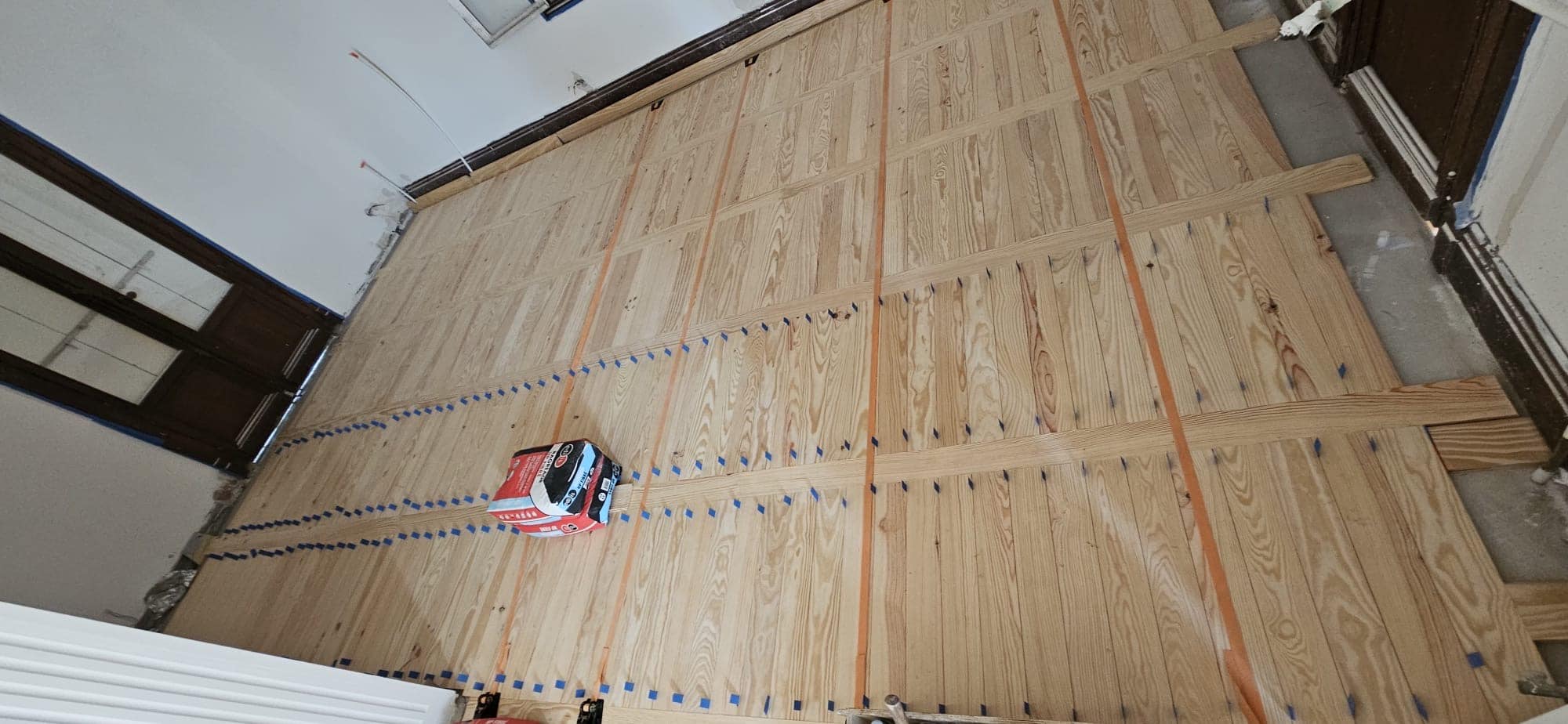 Pose de parquet massif en damier avec sangles de serrage et repères bleus pour une rénovation de sol impeccable.