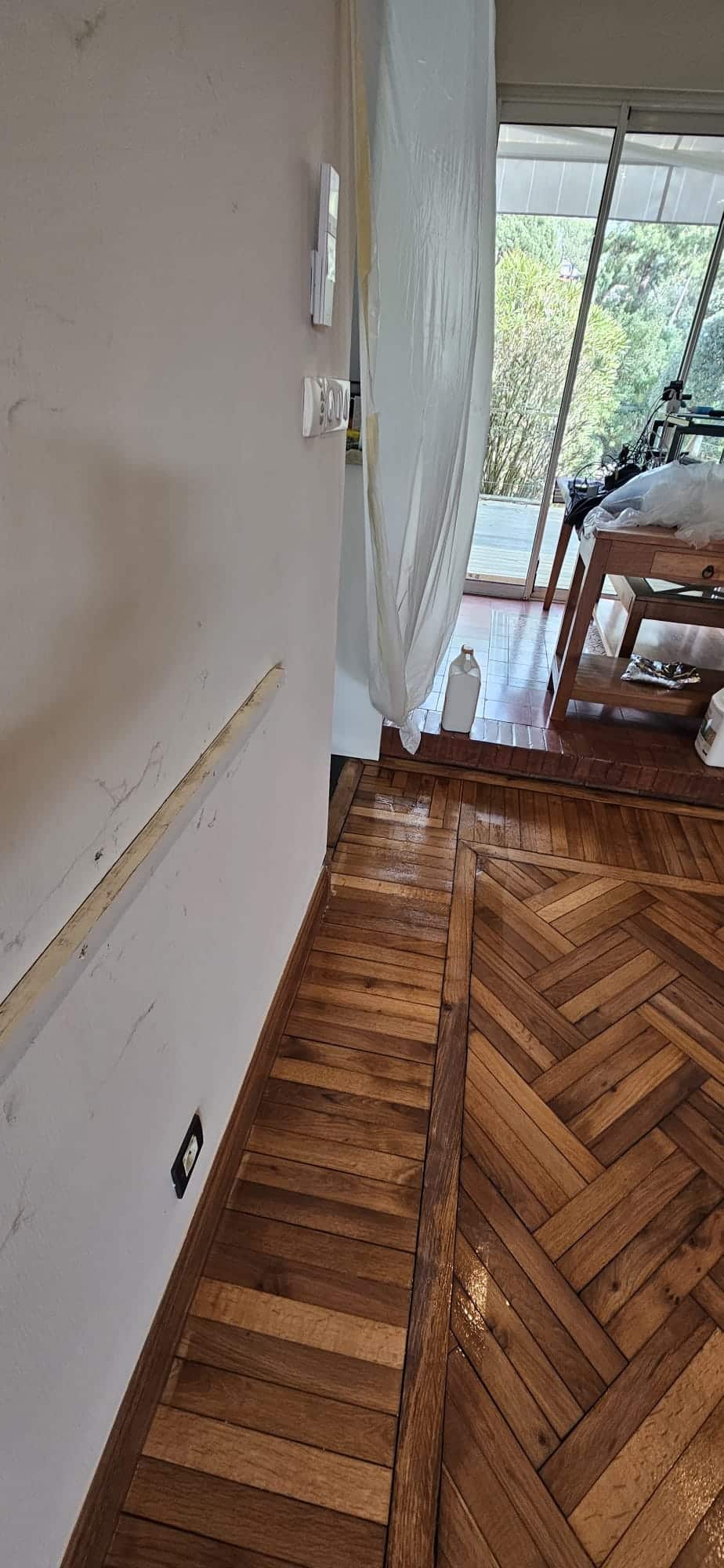Parquet en bois vernis, mur en rénovation avec tasseau et protection plastique devant une baie vitrée.