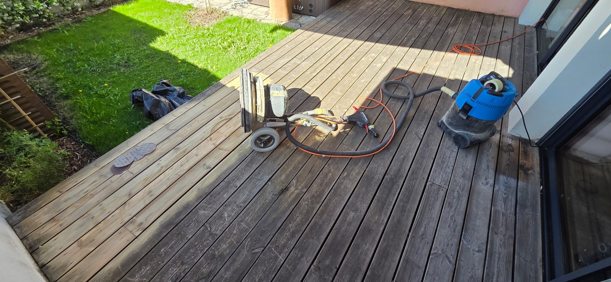 Ponçage d'une terrasse en bois avec ponceuse professionnelle et aspirateur industriel en extérieur.