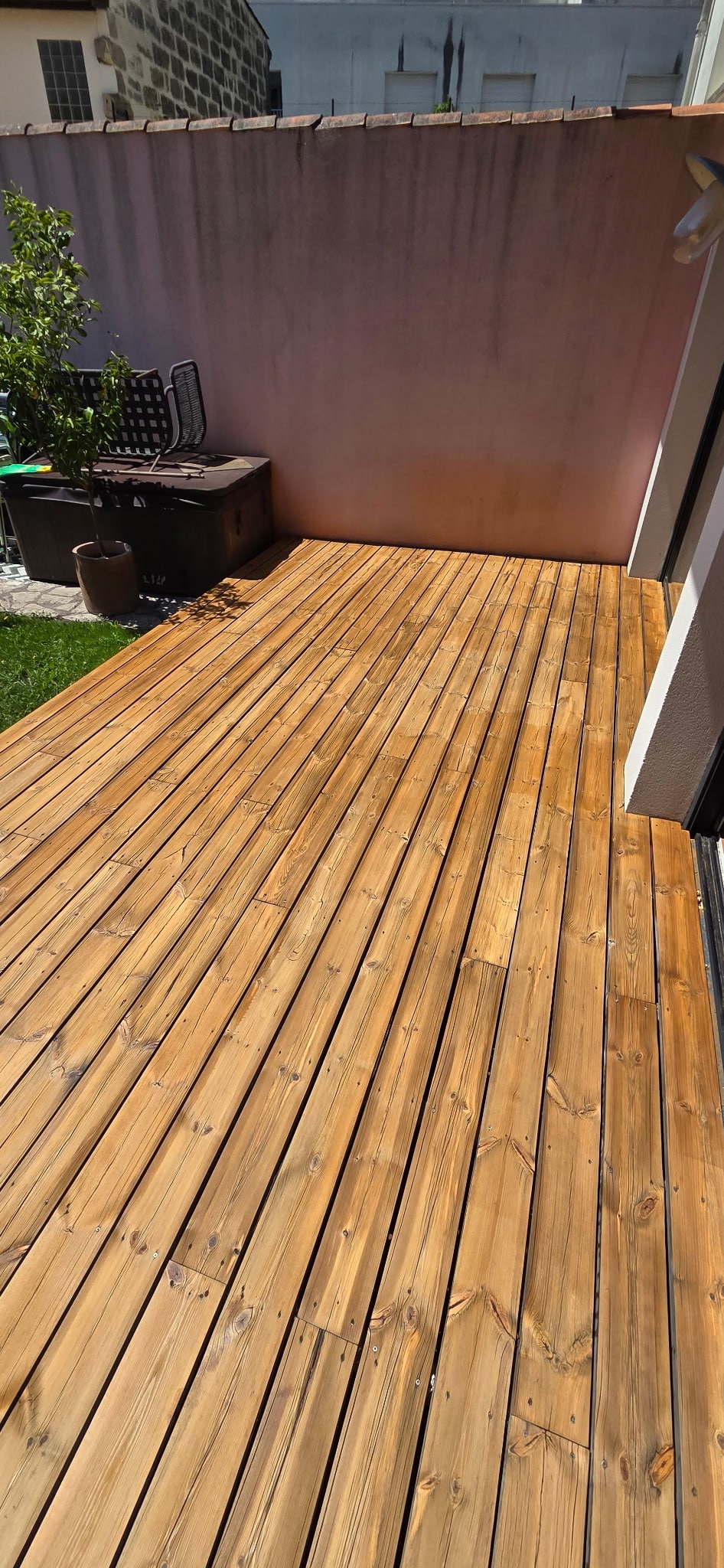 Terrasse en bois clair verni sur un patio extérieur avec mur rose et arbuste.