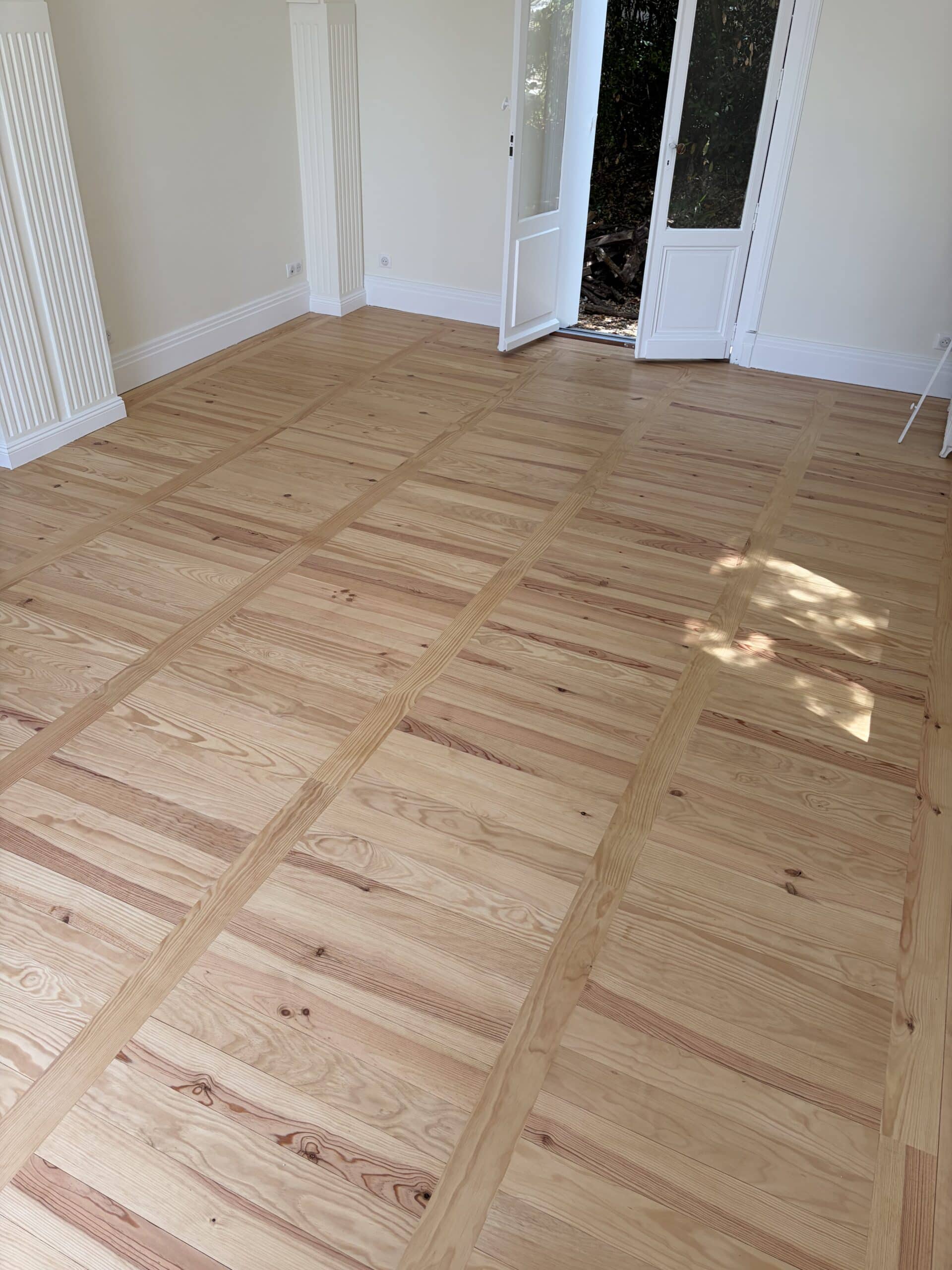 Parquet bois clair à motifs géométriques dans une pièce blanche lumineuse avec double porte ouverte.