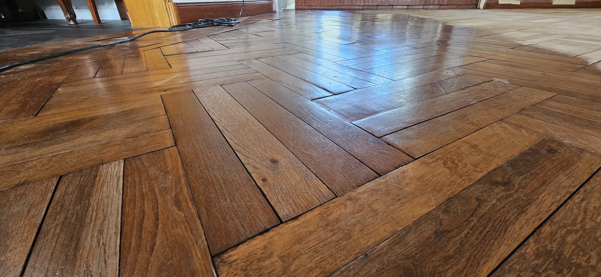 Parquet ancien en bois massif à chevrons, vernis brillant et reflets lumineux dans une pièce en rénovation.