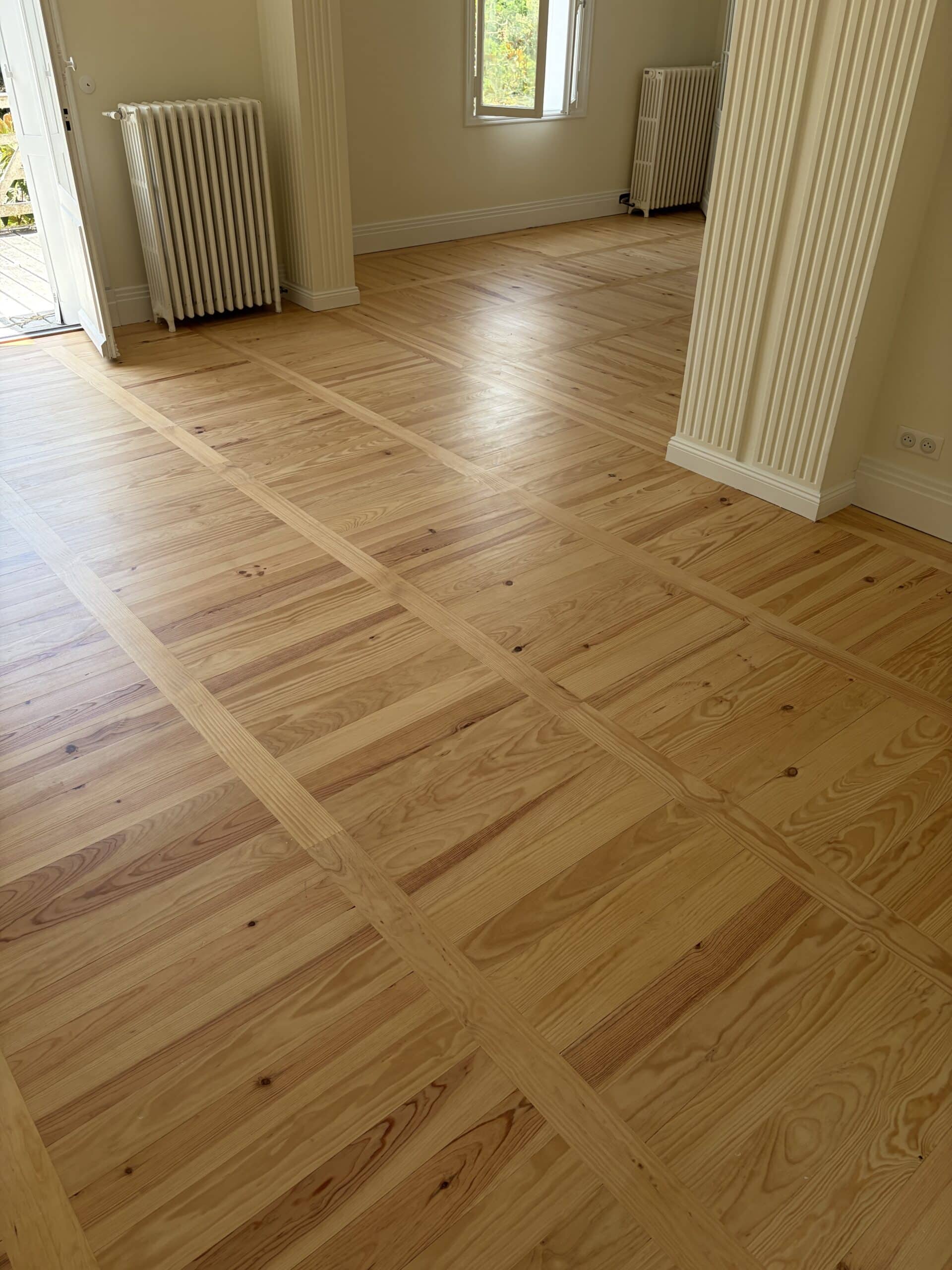 Parquet en bois à motif damier géométrique dans une pièce lumineuse avec colonnes cannelées et radiateurs.