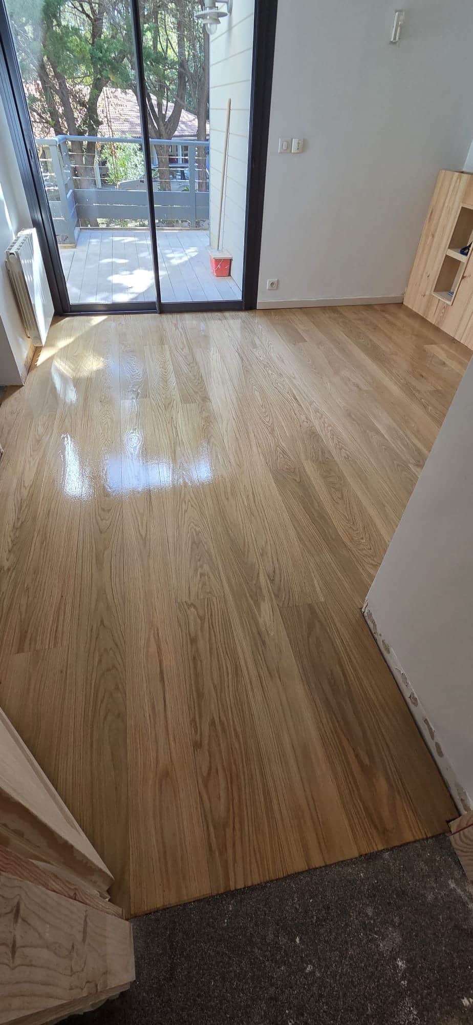 Parquet Chêne Massif : Rénovation et Finition Brillante Parquet chêne massif vitrifié brillant dans une pièce lumineuse ouvrant sur un balcon.